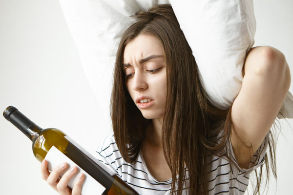 młoda kobieta na kacu z poduszką na głowie wpatruje się w butelkę wina
